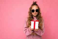 漂亮的快乐年轻的金发女郎卷曲的女人孤立的粉红色的背景墙穿粉红色的体育运动衣服太阳镜持有礼物盒子相机