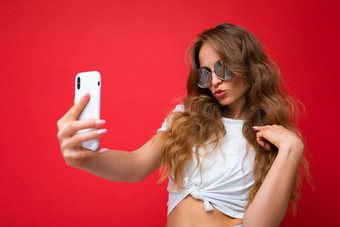 特写镜头照片令人惊异的美丽的年轻的金发女郎女人持有移动电话采取自拍照片智能手机相机穿太阳镜日常时尚的装孤立的色彩斑斓的墙背景设备屏幕