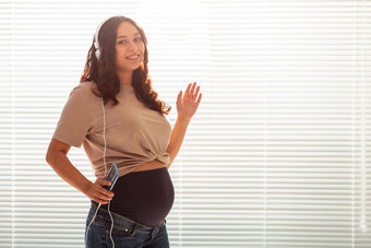 卷发浅黑肤色的女人安抚了怀孕了女人听愉快的经典音乐智能手机耳机概念舒缓的情绪会议婴儿Copyspace