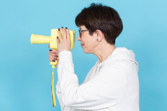 一边视图中年女人短发型毛衣眼镜持有黄色的古董电影相机蓝色的背景概念视频拍摄