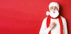 概念圣诞节冬天假期庆祝活动肖像有趣的男人。圣诞老人他持有胡子面具享受一年聚会，派对站红色的背景