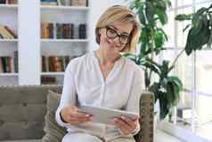 快乐的中间岁的女人坐着沙发电脑平板电脑应用程序屏幕阅读好新闻社会网络购物聊天在线
