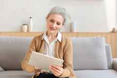 快乐的中间岁的女人坐着沙发电脑平板电脑应用程序屏幕阅读好新闻社会网络购物聊天在线