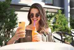 微笑美丽的浅黑肤色的女人女人坐着街夏天咖啡馆自拍移动电话喝汁