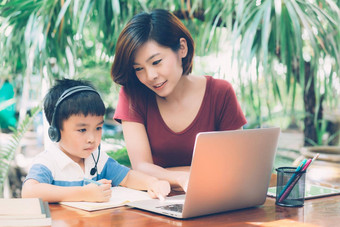 年轻的亚洲妈妈。儿子移动PC电脑研究学习首页男孩写作笔记本家庭作业穿耳机老师妈妈支持孩子教育概念