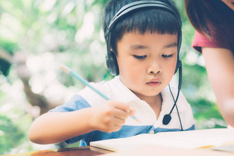 年轻的亚洲妈妈。儿子移动PC电脑研究学习首页男孩写作笔记本家庭作业穿耳机老师妈妈支持孩子教育概念