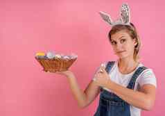 年轻的女人兔子耳朵显示拇指联合包裹持有篮子复活节鸡蛋孤立的粉红色的背景