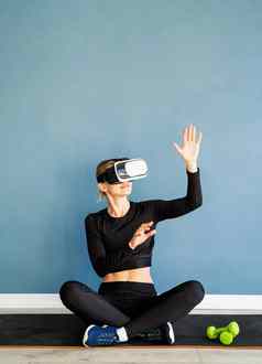 年轻的金发女郎女人体育运动衣服穿虚拟现实眼镜坐着健身席互动菜单