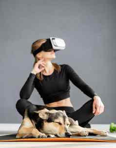 年轻的女人体育运动衣服穿虚拟现实眼镜坐着健身席狗