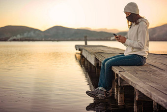 孤独的女人智能手机遵循最新的新闻科维德坐着码头保护面具日落日出正常的活动互联网移动技术冠状病毒