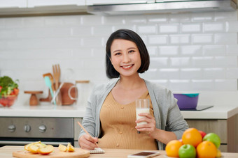 怀孕了女人持有玻璃牛奶准妈妈。摆姿势厨房