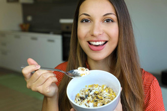关闭美丽的年轻的女人吃斯凯尔酸奶麦片牛奶什锦早餐水果首页相机焦点模型眼睛室内图片健康的早餐概念