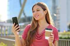 微笑年轻的巴西女人穿粉红色的上衣视频调用智能手机在户外持有外卖咖啡杯