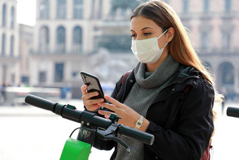 环保运输年轻的业务女人外科手术面具解锁e-scooter移动电话女人应用程序智能手机解锁共享电踏板车城市街