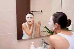 美丽的年轻的亚洲女人应用化妆品面部面具浴室