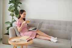 美丽的年轻的亚洲女人移动PC坐着生活房间喝咖啡