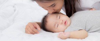 年轻的亚洲妈妈。吻脸颊婴儿女孩温柔的床上卧室妈妈爱新生儿护理妈妈。表达式孩子父女儿家庭概念