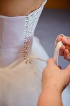 伴娘帮助纤细的新娘用带子束紧婚礼白色衣服钉纽扣精致的花边模式毛茸茸的裙子腰早....新娘准备细节新婚夫妇婚礼一天时刻穿