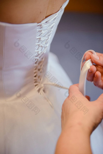 伴娘帮助纤细的新娘用带子束紧婚礼白色衣服钉纽扣精致的花边模式毛茸茸的裙子腰早....新娘准备细节新婚夫妇婚礼一天时刻穿