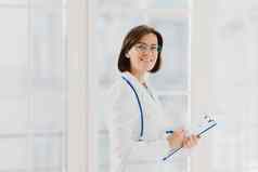 水平拍摄女医生填满医疗形式剪贴板站室内穿轮眼镜白色礼服听诊器一般医生写笔记咨询病人