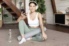 微笑亚洲女孩耳机穿体育运动服装听音乐持有移动电话锻炼首页坐着生活房间
