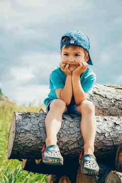 男孩蓝色的棒球帽牛仔布短裤坐在日志深思熟虑