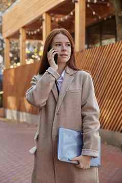高加索人女人时尚的米色外套调用智能手机持有移动PC