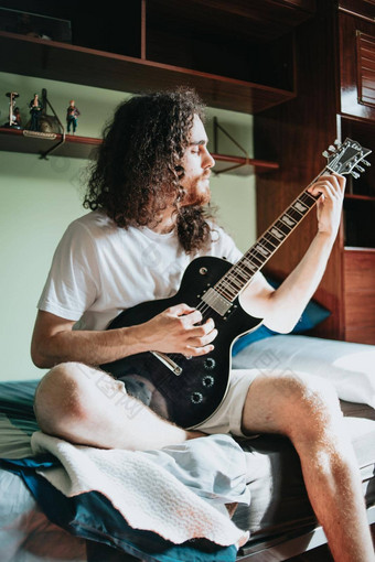 年轻的男人。长头发玩电吉他房间白色衬衫坐着舒适的床上卧室思考<strong>音乐教育</strong>概念图像