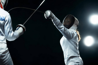 年轻的击剑运动员穿面具白色击剑服装持有剑黑色的背景灯