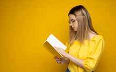 美丽的聪明的年轻的女孩持有阅读书孤立的黄色的背景肖像有吸引力的女人黄色的上衣穿眼镜阅读书教育研究知识