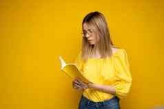 美丽的聪明的年轻的女孩持有阅读书孤立的黄色的背景肖像有吸引力的女人黄色的上衣穿眼镜阅读书教育研究知识