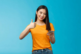 满意可爱的亚洲浅黑肤色的女人持有智能手机显示翘拇指很高兴推荐太棒了小工具给批准好应用程序同意朋友<strong>发布</strong>好博客文章站蓝色的<strong>背景</strong>