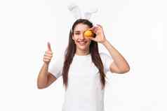 肖像满意快乐年轻的女人兔子耳朵持有彩色的鸡蛋眼睛显示竖起大拇指绘画复活节假期聚会，派对庆祝传统的事件白色背景