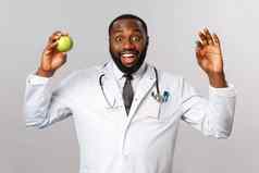 医疗保健医学健康的生活方式概念热情的非裔美国人医生建议保持健康的吃维生素水果持有苹果药片站灰色背景
