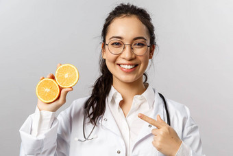 特写镜头快乐的有吸引力的女医生白色外套眼镜指出橙色建议吃健康的食物水果蔬菜保持安全护理健康灰色背景