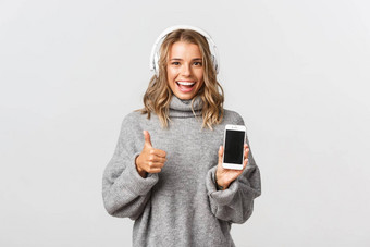 图像满意金发碧眼的女孩快乐显示竖起大拇指听音乐耳机演示智能手机屏幕白色背景