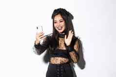 肖像美丽的时尚的亚洲女人哥特花边衣服挥舞着手智能手机相机视频调用站白色背景