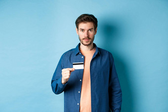 英俊的年轻的男人。胡子穿休闲衣服显示塑料信贷卡相机蓝色的背景