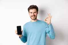 电子商务购物概念英俊的有胡子的的家伙显示空移动屏幕标志赞扬好应用程序站白色背景
