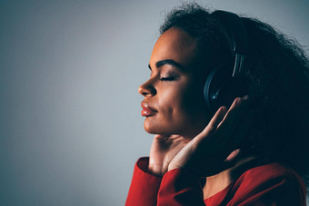<strong>快乐音乐</strong>年轻的非裔美国人女孩听最喜欢的跟踪耳机穿红色的夹克黑色的前孤立的灰色背景在情感上移动有趣的冷健美的图像