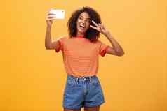 女孩使视频视频博客品牌智能手机发布互联网著名的站橙色背景摆姿势自拍盯着小工具屏幕显示和平胜利手势