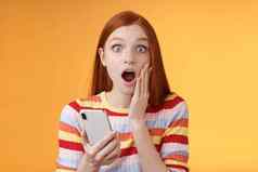 有关震惊了感情的红色头发的人女孩找到惊人的新闻相机下降下巴喘气深刻的印象触摸脸颊持有智能手机完成阅读令人印象深刻的令人不安的故事橙色背景
