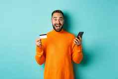 在线购物惊讶男人。持有移动电话信贷卡支付互联网商店站光蓝色的背景