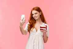 诱人的高加索人生日女孩红色的长头发白色衣服采取自拍帖子互联网社会媒体生日聚会，派对持有智能手机和平美味的蛋糕粉红色的背景