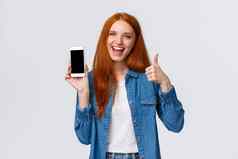 女孩显示朋友很酷的移动游戏有吸引力的快乐的红色头发的人女人持有智能手机介绍应用程序电话应用程序使翘拇指微笑批准推荐