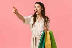女人美妙的衣服商店manequin有吸引力的快乐的年轻的女衣服持有购物袋指出手指横盘整理喘气惊讶站粉红色的背景