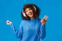 快乐的无忧无虑的可爱的非裔美国人女模型冬天毛衣跳舞节奏音乐听跟踪首歌应用程序圣诞节播放列表穿耳机持有智能手机