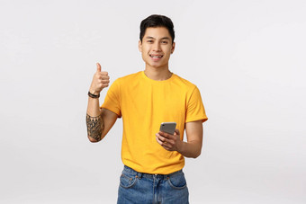 太棒了内容伟大的在线购物网站快乐的亚洲赶时髦的人的家伙纹身穿时尚的黄色的t恤促进智能手机应用程序移动内容持有智能手机显示拇指