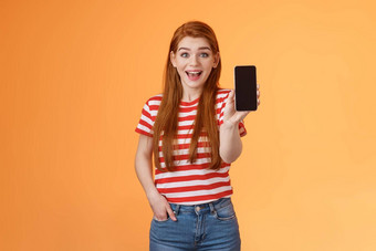 兴奋可爱的即将离任的红色头发的人女深刻的印象显示应用程序持有智能手机介绍小工具功能微笑惊讶热情的应用程序吹牛游戏分数橙色背景