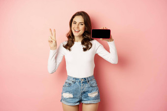 时尚的年轻的女人<strong>介绍公司</strong>网站移动应用程序显示智能手机水平和平标志微笑快乐站粉红色的背景
